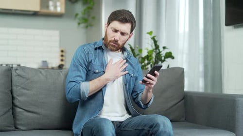 Man having heart attack sitting alone at home on the sofa. Mature bearded Caucasian male holding