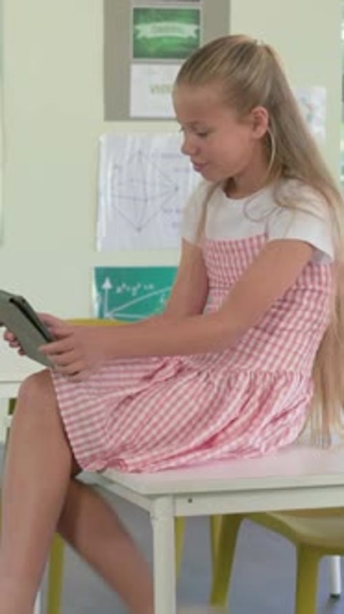 Vertical video: Sitting girl in pink dress holding tablet on classroom table, reading with earbuds