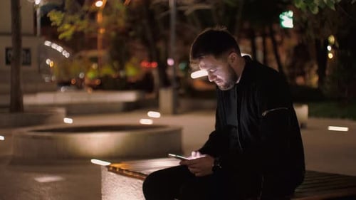 Night time medium shot of a man sitting on a bench in a park, writing-texting on a smartphone with t