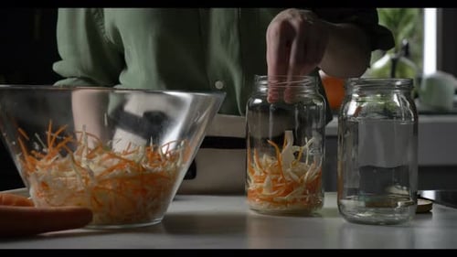 white housewife places carrots and cabbage for fermentation into a glass jar at home on the kitchen