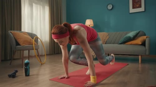 Redhaired Young Woman in Pink Sportswear is Warming Up Stretching on a Sports Mat in the Living Room