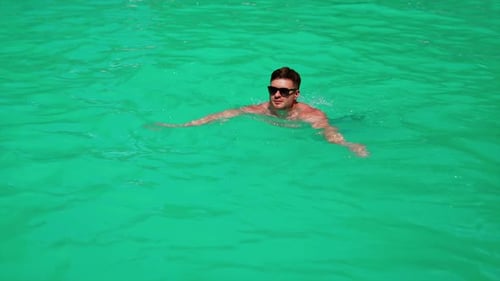 Caucasian man swimming in the pool. Male wearing sunglasses swims in azure water in the sun.