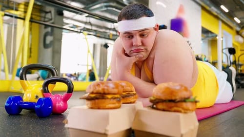 Young Adult Man Lying in Gym Looking at Burgers