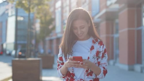 Smiling Curly Woman Wearing Trendy Sunglasses Walks Down the Central City Street and Uses Her Phone
