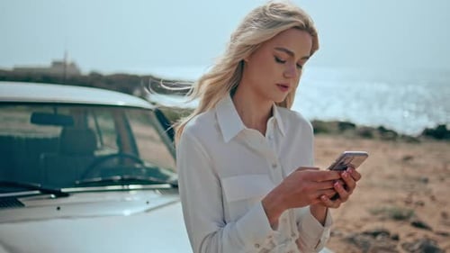 Gorgeous blonde woman texting on smartphone leaning at retro car on sunny beach