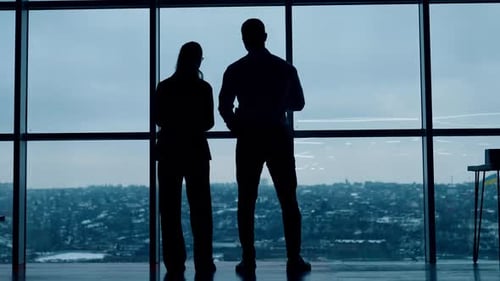 Dark silhouettes of a couple standing looking at the window. Colleagues in office look at cityscape.