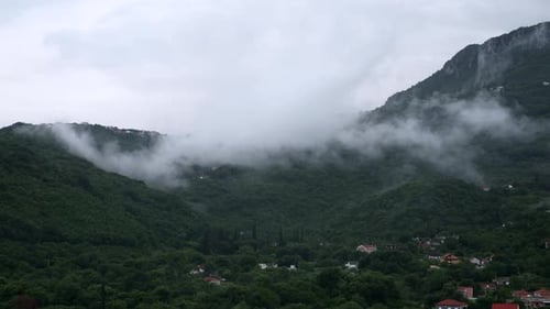 Kleines Dorf in hohen Bergen, mit Bäumen bewachsen und mit niedrigen Regenwolken bedeckt.