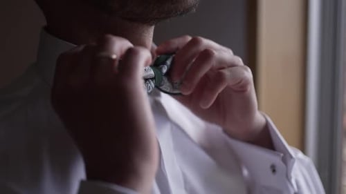 Man adjusting his patterned bow tie, wearing cufflinks and a wedding ring, in a close-up view