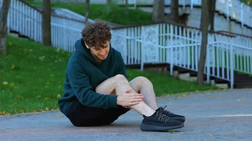 Sportsman Sitting on Ground and Stretching His Knee Leg Muscles in Green Park Before Outdoor