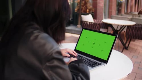Young Girl Watching on a Laptop with a Mockup Green Screen Woman in a Black Jacket is Sitting at the