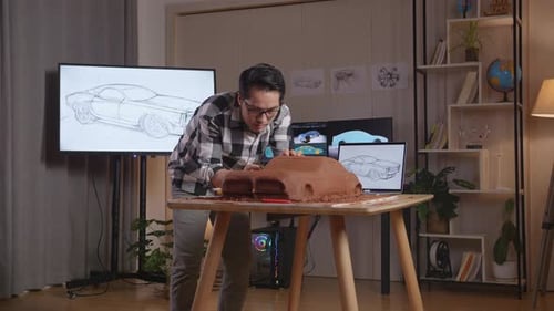Asian Man Automotive Designer Looking At A Laptop And Using Rake Or Wire To Smooth Out Car Clay