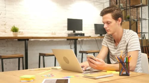 Young Man Using Phone and Laptop in Office