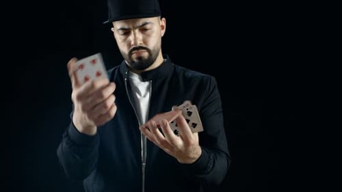 Professional Street Magician in a Cap Performs Sleight of Hand Card Tricks. Background is Black.