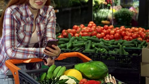 Smiling Woman with Produce Shopping Cart Using Phone