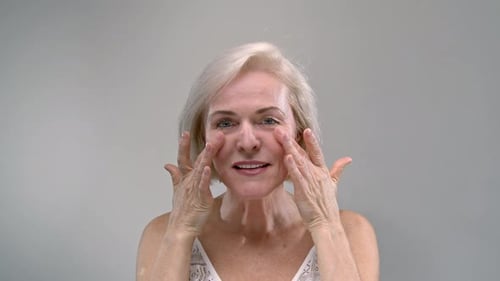 Old Woman Touching Facial Skin and Head Smiles Senior Female in the White Studio Beauty Concept