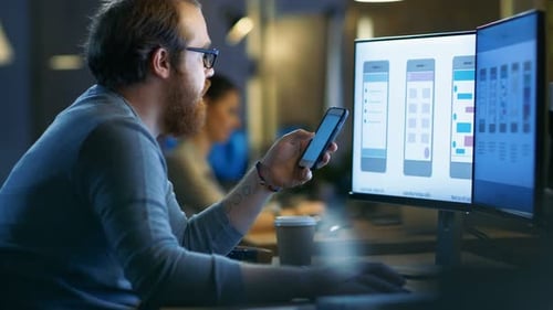 Male Mobile Application Designer Tests New Features on His Smartphone, He Works on a Personal Compu