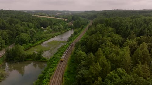 Aerial View of a Yellow Train Traveling on a Railway Track Next to a River in Belgium Surrounded By