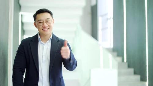 Smiling asian businessman showing thumb up, like gesture, standing in modern office building.