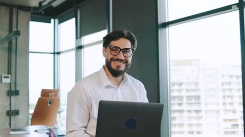 Smiling Businessman Working on Laptop in Modern Office During Daylight with Large Windows and Urban