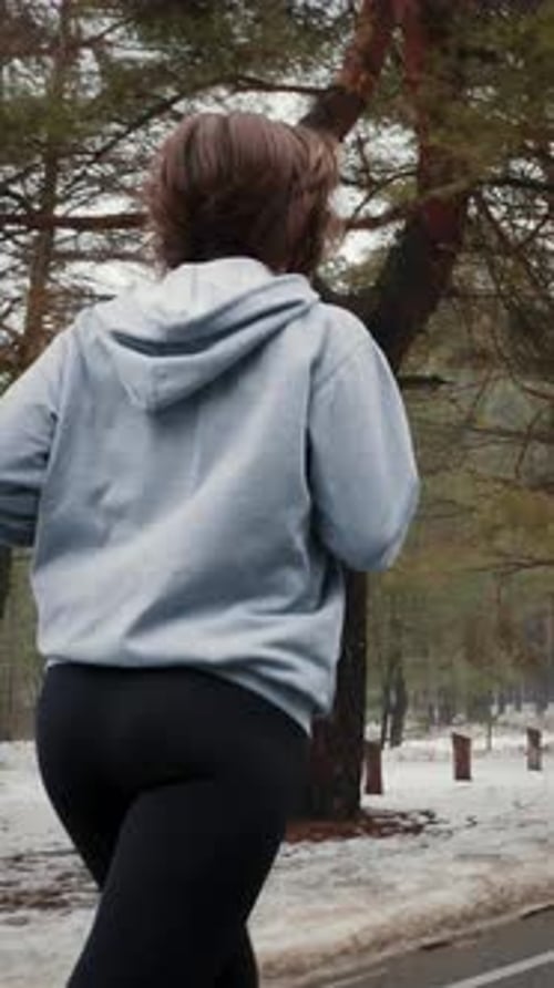 Female in hoodie and leggins jogging in winter park, back shot. Vertical video