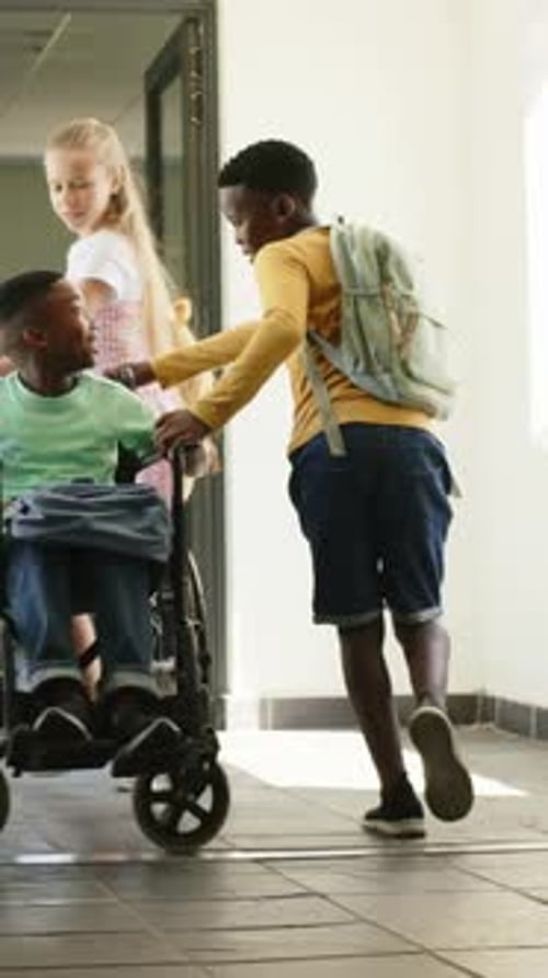 Vertical video: Holding door, yellow helper helping friend in wheelchair into school hall with pack