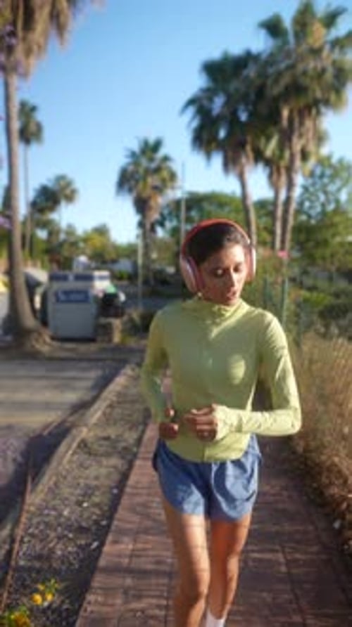 Woman in Sportswear Jogging Outdoors Listening to Music with Headphones in Greenery on a Sunny Day