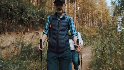 Couple of Pensioners with Backpacks and Hiking Sticks Walking on a Narrow Forest Trail in the Dense