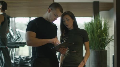 Athletic man and woman are using a tablet next to treadmills in a sport gym.