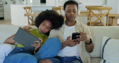 African American couple sitting on couch using smartphone and tablet, man wearing white headphones