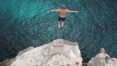 Overhead slow motion aerial: Man jumps off rocky cliff into green sea