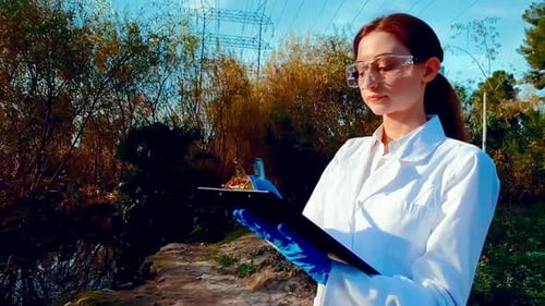 A young woman scientist at a creek, wearing protective eyewear and a lab coat, taking notes on a cli