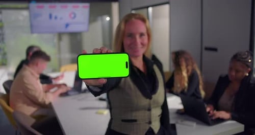 Smiling businesswoman holding smartphone with green screen in modern office, colleagues working