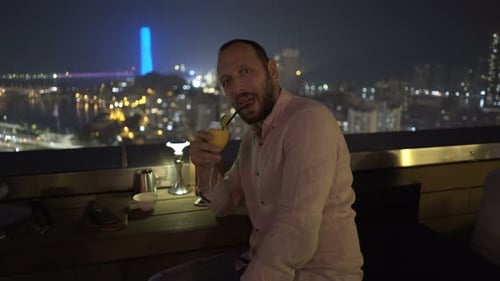 Happy young man raising toast with cocktail drink on luxury rooftop terrace at night