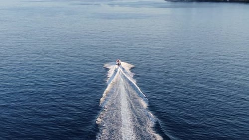 Aerial drone follows a person on a jet ski speeding across the open blue sea, leaving a large white