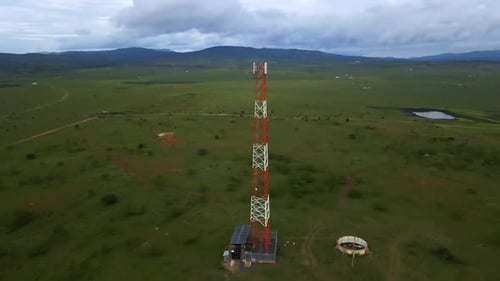 An aerial view of a cell tower standing tall in a vast, undulating green field, surrounded by a few