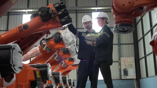 Une technicienne programme un bras robotique avec une tablette numérique et un robot d'assemblage dans une usine.