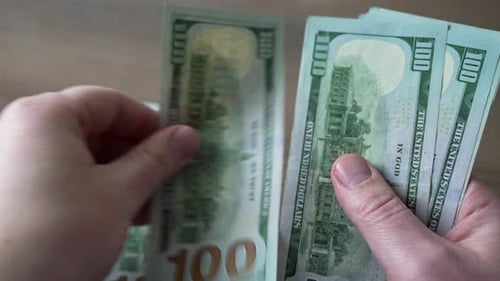 Mãos masculinas contando notas de cem dólares em close-up de uma mesa de madeira.