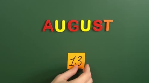 Hand sticking a sticker 13 August calendar day on school board. 13 date of august. Thirteenth day