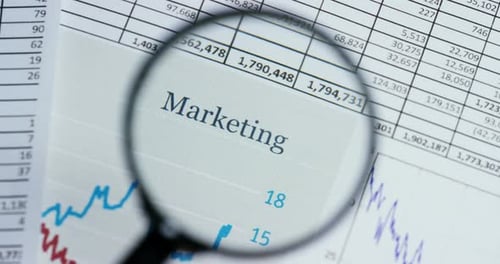 A hand with a magnifying glass passes over a financial newspaper and financial bag with a business