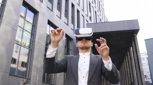 Man in stylish wear which working on virtual screen in augmented reality goggles near city building,