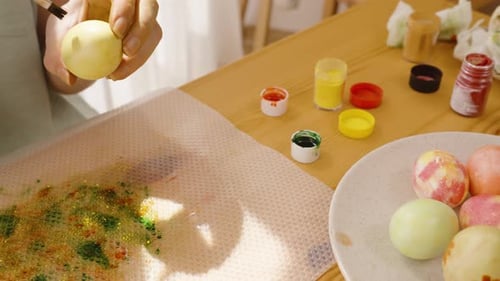 Woman decorating an Easter egg with green dots in a bright, sunny room.