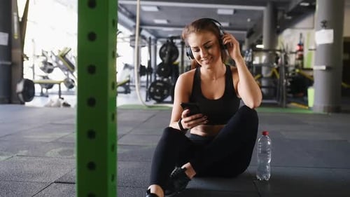 Young girl in sportive clothes is in the gym at daytime taking break and listening to the music