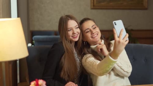 Good-looking young female friends in cafe are smiling and taking selfies on smartphone. Side view of