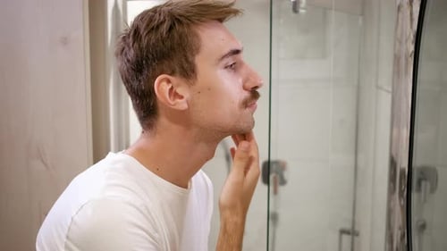 Junger Mann mit Schnurrbart schaut sich seine Haut im Badezimmerspiegel an.