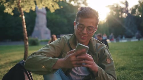 Portrait Young Man with Glasses Using Smartphone Listen Music on Headphone Sitting in Park Smile