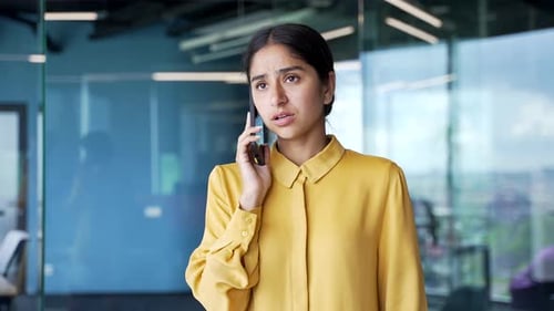 Upset young businesswoman standing in modern office talking on smartphone. Unhappy female employee