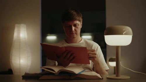Focused Man Reading Paper Sitting at Table Book Under Soft Glow of Lamp in Dimly Lit Room at Night