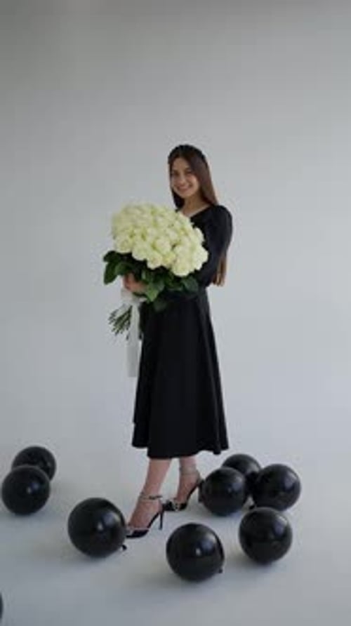 Young Woman in Black Dress Holding Bouquet of White Roses Standing Among Black Balloons in Studio