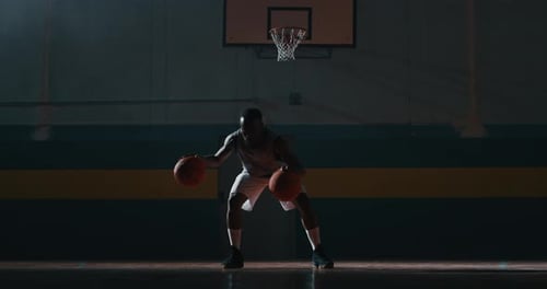 Basketball Player Dribbles Two Balls in Dark Gymnasium