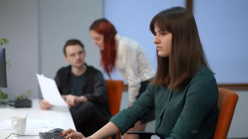 Side View Concentrated Young Woman Working in Office with Blurred Colleagues Talking at Background